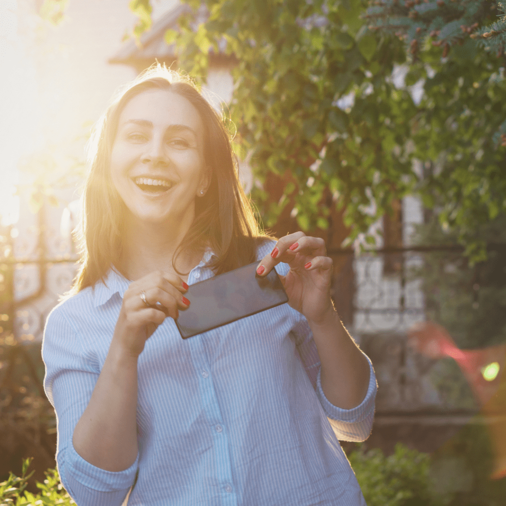 Uśmiechnięta kobieta trzymająca smartfon w promieniach zachodzącego słońca na tle zieleni.
